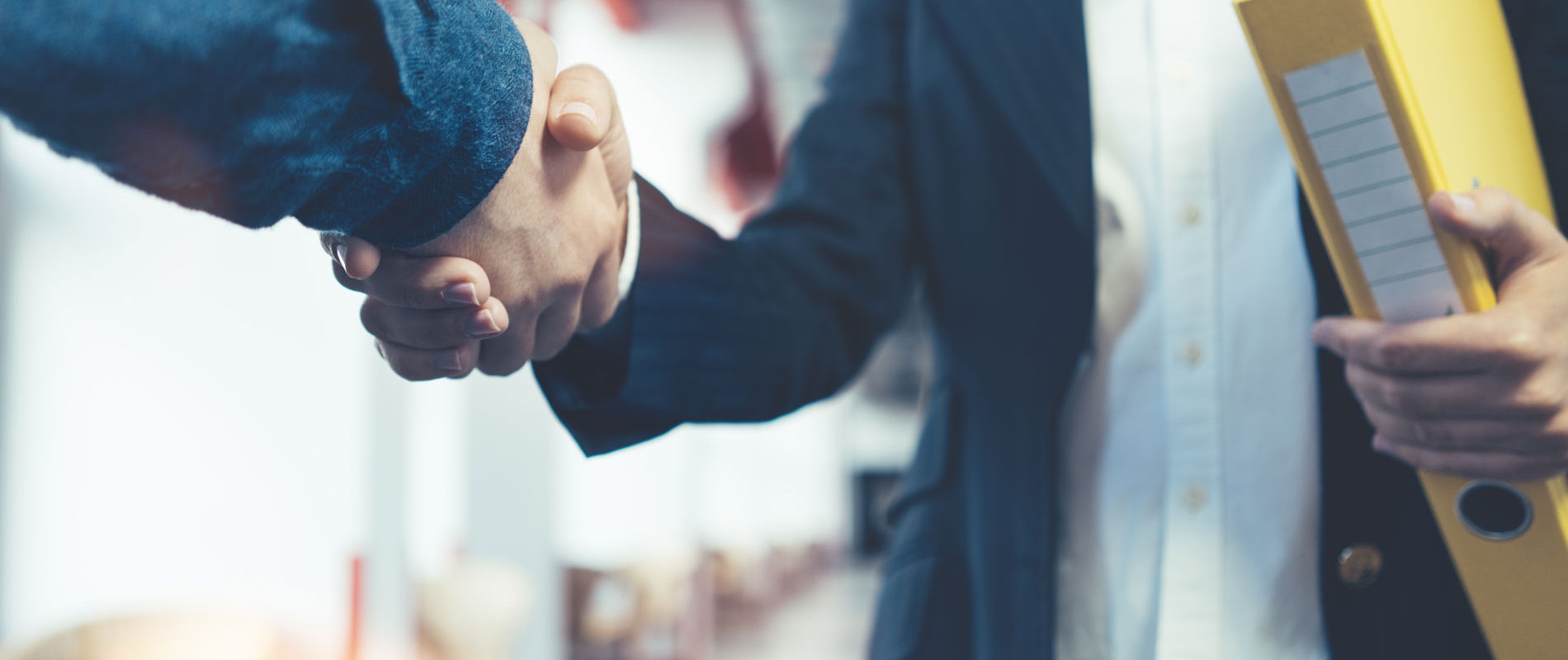 Zwei Personen schütteln sich die Hand. Eine Person hält einen gelben Ordner. Bürohintergrund unscharf.