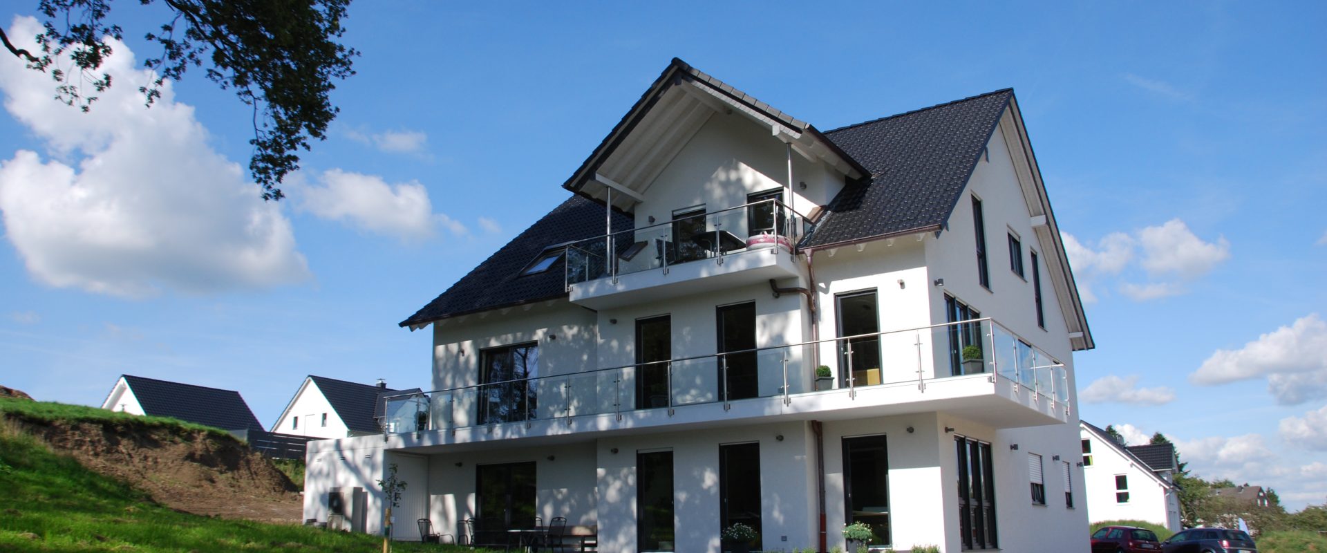 Ein modernes, weißes Zweifamilienhaus mit Balkon und großzügigem Garten unter blauem Himmel.