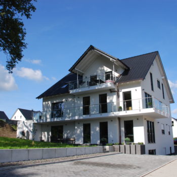 Ein modernes, weißes Zweifamilienhaus mit Balkon und großzügigem Garten unter blauem Himmel.